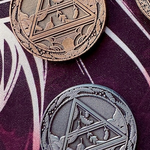 May include: Close-up of two round, metallic coins with intricate designs. The coins feature a central triangle with leaf and feather details, surrounded by a decorative border. One coin is bronze-colored, the other is silver-colored.