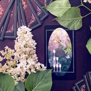 K&ouml;nnte beinhalten: Eine Tarotkarte mit schwarzem Hintergrund und einem wei&szlig;en ovalen Rahmen. Die Karte zeigt einen Stapel von f&uuml;nf Kelchen in einer Blumenumgebung. Die Karte ist von gr&uuml;nen Bl&auml;ttern und wei&szlig;en Bl&uuml;ten umgeben.