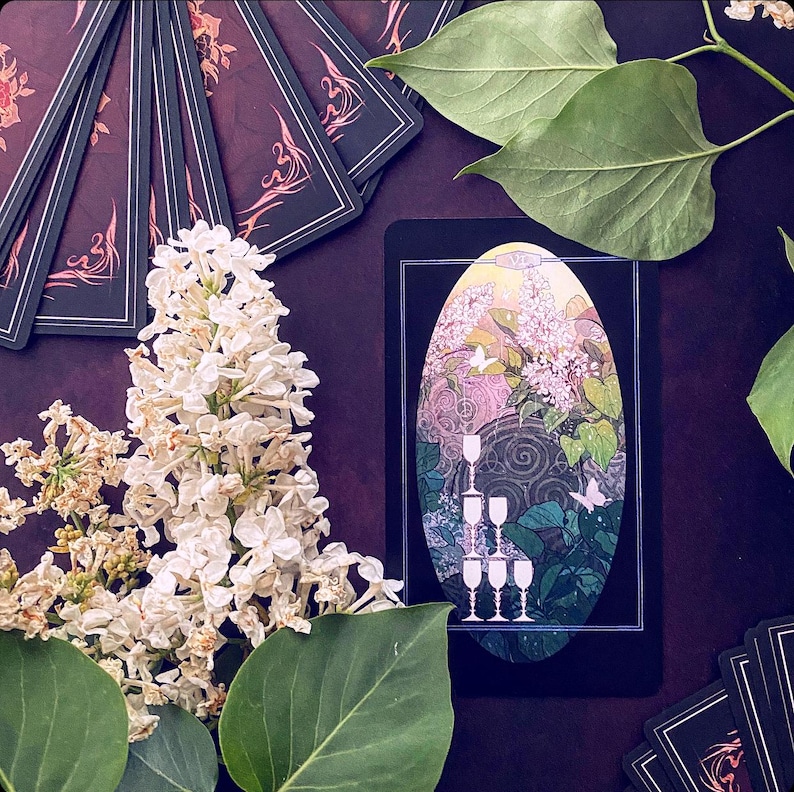 K&ouml;nnte beinhalten: Eine Tarotkarte mit schwarzem Hintergrund und einem wei&szlig;en ovalen Rahmen. Die Karte zeigt einen Stapel von f&uuml;nf Kelchen in einer Blumenumgebung. Die Karte ist von gr&uuml;nen Bl&auml;ttern und wei&szlig;en Bl&uuml;ten umgeben.
