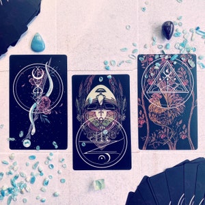 May include: Three tarot cards with black backgrounds and intricate designs. The first card features a black and white illustration of a creature with a crown and a geometric pattern. The second card features a black and white illustration of a creature with a crown and a geometric pattern. The third card features a black and white illustration of a rooster with a geometric pattern.