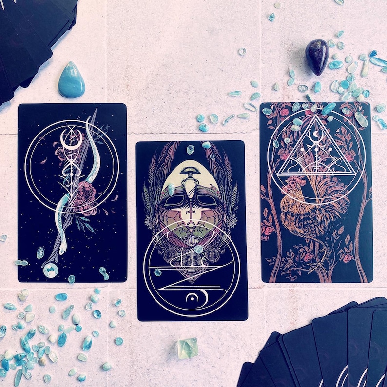 May include: Three tarot cards with black backgrounds and intricate designs. The first card features a black and white illustration of a creature with a crown and a geometric pattern. The second card features a black and white illustration of a creature with a crown and a geometric pattern. The third card features a black and white illustration of a rooster with a geometric pattern.