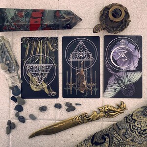 May include: Three tarot cards with black backgrounds and white designs. The card on the left has a white triangle with a circle inside and a white design of a creature with wings. The card in the middle has a white triangle with a circle inside and a white design of a creature with wings. The card on the right has a white circle with a purple flower inside and a white design of a creature with wings.