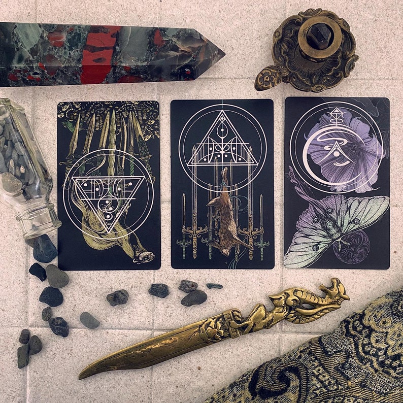 May include: Three tarot cards with black backgrounds and white designs. The card on the left has a white triangle with a circle inside and a white design of a creature with wings. The card in the middle has a white triangle with a circle inside and a white design of a creature with wings. The card on the right has a white circle with a purple flower inside and a white design of a creature with wings.