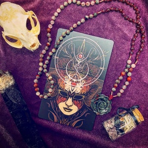 May include: A black and gold tarot card with a detailed illustration of a masked figure with horns. The card is surrounded by a beaded necklace with green, red, and brown stones. The necklace is draped over a purple velvet surface.