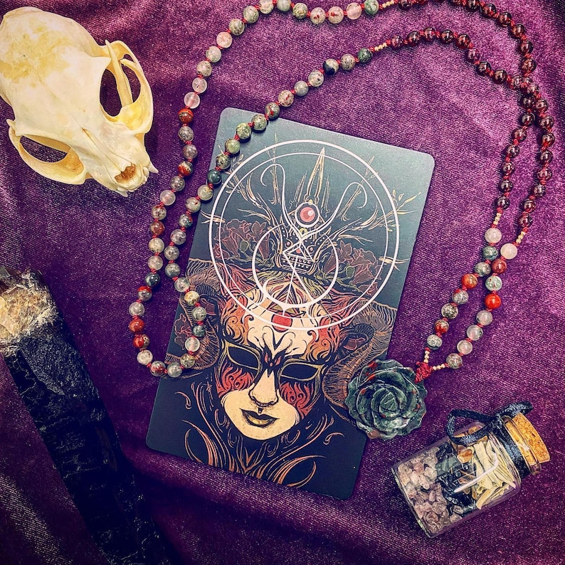 May include: A black and gold tarot card with a detailed illustration of a masked figure with horns. The card is surrounded by a beaded necklace with green, red, and brown stones. The necklace is draped over a purple velvet surface.