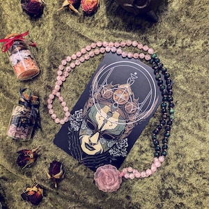 May include: A pink and green beaded necklace with a black card featuring a detailed illustration of a masked figure with a geometric design. The card has the text "The Fool" printed on it.