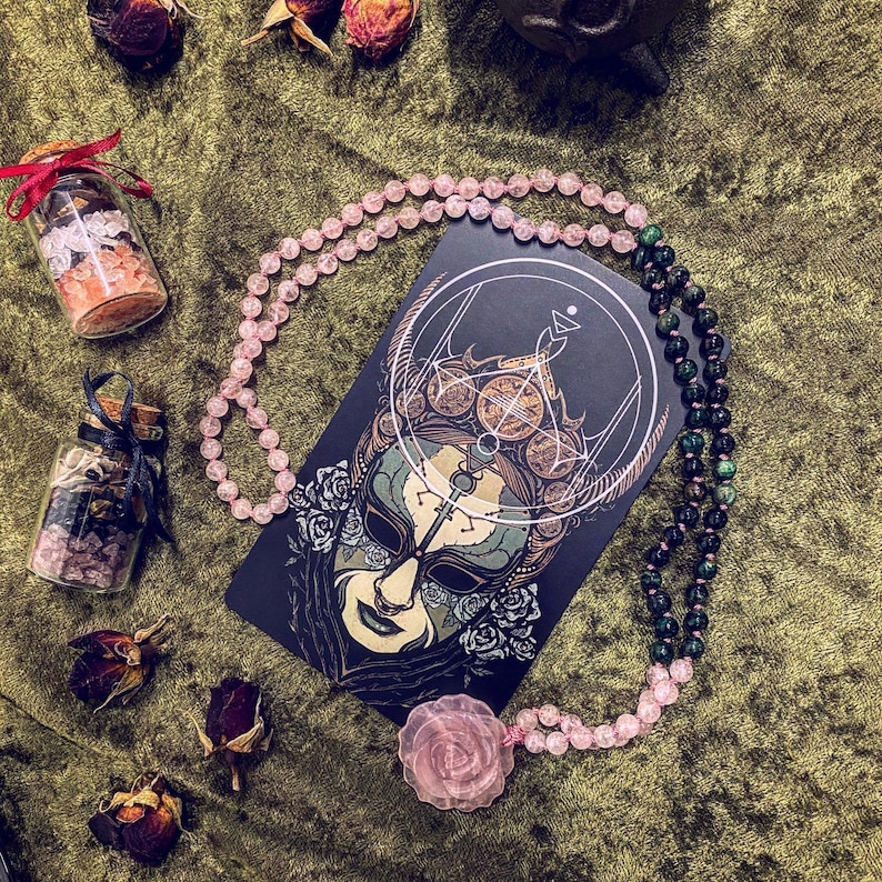 May include: A pink and green beaded necklace with a black card featuring a detailed illustration of a masked figure with a geometric design. The card has the text "The Fool" printed on it.