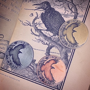 May include: Three round metal coins with a raven silhouette design. The coins are in different colors: silver, gold, and copper. The coins are on a book with a black and white illustration of a raven.