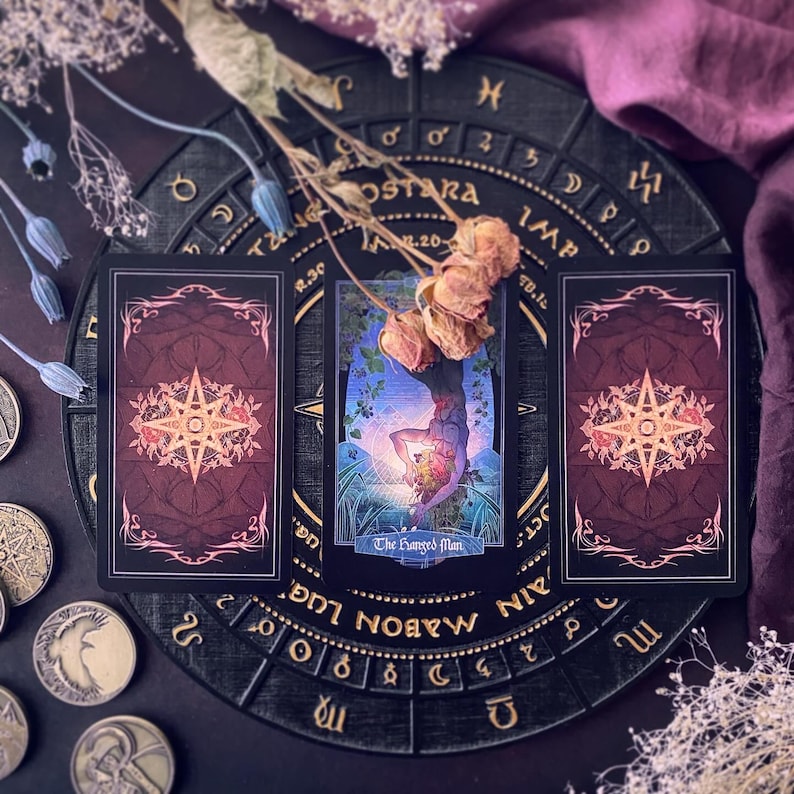 K&ouml;nnte beinhalten: Drei Tarotkarten auf einem schwarzen runden Brett mit goldenen Buchstaben. Die Karten sind schwarz mit goldenen Verzierungen und zeigen ein Sternmuster. Die mittlere Karte zeigt eine Person, die kopf&uuml;ber in einem Baum h&auml;ngt, mit dem Text "The Hanged Man".