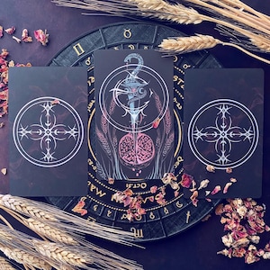 May include: Three tarot cards with black backgrounds and white designs. The center card features a blue and silver dagger with a skull and crossbones design. The card is surrounded by wheat stalks. The other two cards feature a white cross with four points.