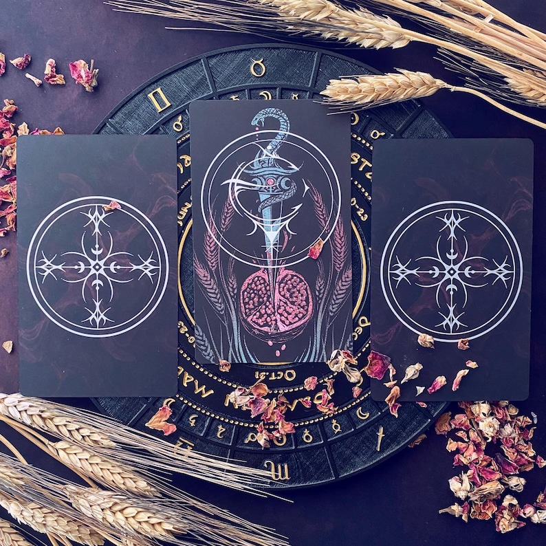 May include: Three tarot cards with black backgrounds and white designs. The center card features a blue and silver dagger with a skull and crossbones design. The card is surrounded by wheat stalks. The other two cards feature a white cross with four points.