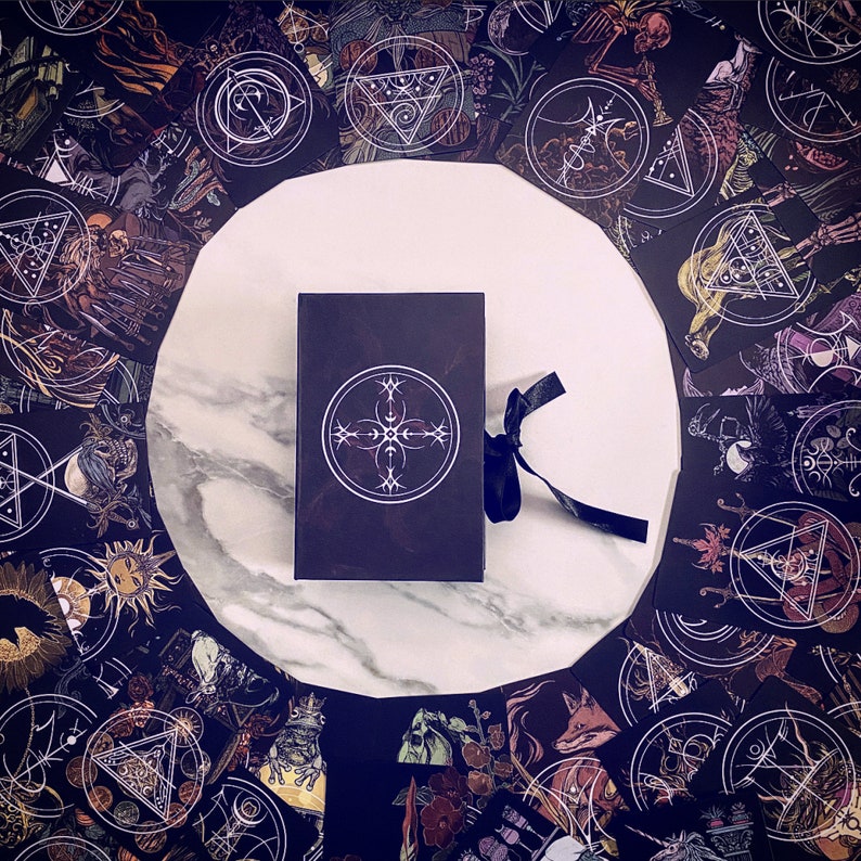 May include: A black box with a white geometric design on the front, surrounded by a variety of cards with intricate designs. The cards feature a dark background with white and gold accents.