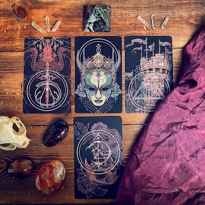 May include: Five tarot cards with intricate designs. The cards feature a dark background with gold accents and illustrations of a masked figure, a castle, and a sword. The cards are laid out on a wooden surface.