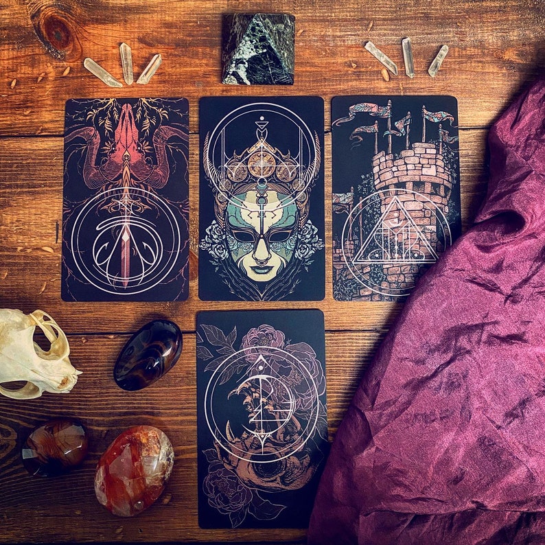 May include: Five tarot cards with intricate designs. The cards feature a dark background with gold accents and illustrations of a masked figure, a castle, and a sword. The cards are laid out on a wooden surface.