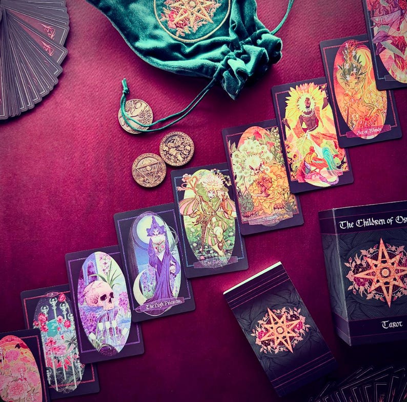 K&ouml;nnte beinhalten: Ein Tarot-Deck mit einem gr&uuml;nen Samtbeutel und M&uuml;nzen. Die Karten zeigen Illustrationen von Feen, Blumen und anderen mystischen Bildern. Die Schachtel f&uuml;r das Deck tr&auml;gt den Text "The Children of the Gods Tarot" auf der Vorderseite.