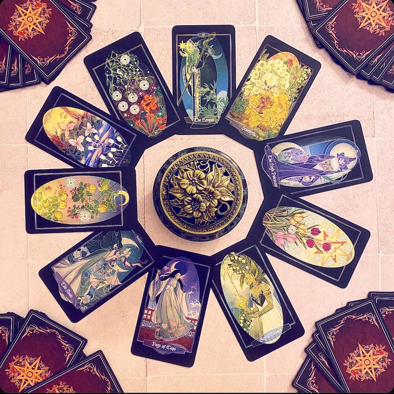 K&ouml;nnte beinhalten: Eine Tarotkarten-Lesespreizung mit 10 Karten, die in einem Kreis um einen dekorativen Metall-R&auml;ucherofen angeordnet sind. Die Karten sind schwarz mit goldenen Akzenten und zeigen Illustrationen von Blumen, Menschen und anderen Symbolen. Die Karten beinhalten "Die Kaiserin", "Die Hohepriesterin", "Der Page der Kelche" und "Der Stern".