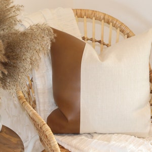 Puede incluir: Un cojín blanco y marrón con un panel de imitación de cuero marrón en un lado. El cojín está sentado en una silla de mimbre con una manta blanca.