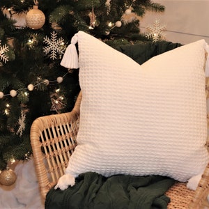 Puede incluir: Cojín blanco con borlas en una silla de mimbre delante de un árbol de Navidad decorado con pompones blancos y copos de nieve.