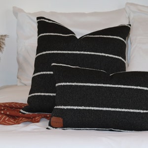 Puede incluir: Dos almohadas de tiro a rayas blancas y negras sobre una cama blanca. La almohada más grande está detrás de la más pequeña. Ambas almohadas tienen una etiqueta de cuero marrón.