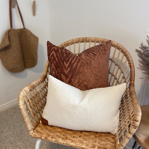 Puede incluir: Una silla de mimbre con patas blancas y dos almohadas. La silla tiene una almohada con estampado marrón y blanco y una almohada blanca. La silla está sobre una alfombra beige.