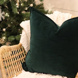 Puede incluir: Una almohada de terciopelo verde oscuro se sienta en una silla de mimbre con una manta blanca. La silla está frente a un árbol de Navidad decorado con copos de nieve blancos y pompones blancos.