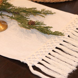 May include: White table runner with a knotted fringe and a sprig of greenery with a pine cone.