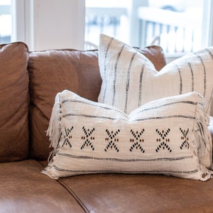 Puede incluir: Dos cojines blancos con patrones geométricos negros y marrones y flecos en un sofá de cuero marrón.