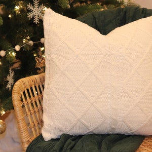 Puede incluir: Un cojín de punto color crema con un patrón de diamantes descansa sobre una silla de mimbre. El cojín está colocado sobre una manta verde oscuro. Un árbol de Navidad con adornos de copos de nieve blancos y luces está al fondo.