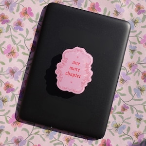 Op de afbeelding: Een zwart elektronisch apparaat met een roze sticker met de tekst "one more chapter" in rode letters. De sticker heeft een decoratieve rand. Het apparaat ligt op een bloemig oppervlak met roze, paarse en groene bloemen.