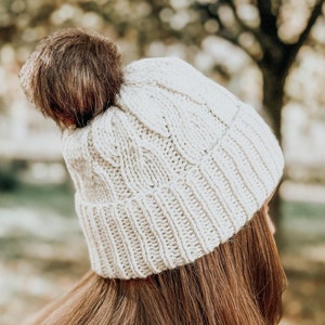 Op de afbeelding: Een witte gebreide beanie met een bruine pompon bovenop. De beanie wordt gedragen door een persoon met lang bruin haar.