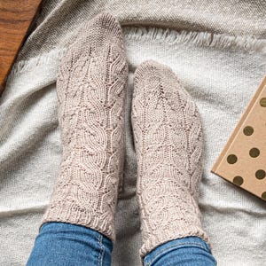 May include: A pair of beige knitted socks with a textured leaf pattern. The socks are worn over blue jeans. A wooden tray with a candle and a gold polka dot gift box are in the background.