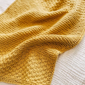 May include: A soft, yellow knitted throw blanket with a textured weave. The blanket is folded and draped over a white surface.