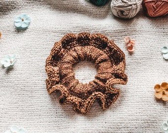 May include: A brown knitted hair scrunchie with a ruffled edge. The scrunchie is lying on a white knitted fabric background.