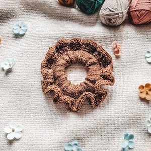 May include: A brown knitted hair scrunchie with a ruffled edge. The scrunchie is lying on a white knitted fabric background.