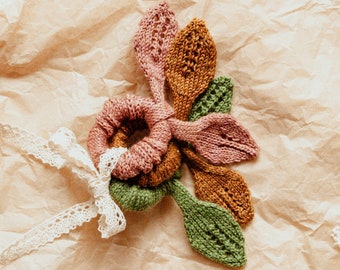May include: A set of four knitted hair scrunchies in shades of pink, green, and brown. The scrunchies are arranged in a flower shape on a brown paper background.