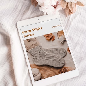 May include: A digital tablet displaying a knitting pattern for Cozy Wafer Socks by whileberry. The pattern features a close-up of the knitted socks on a brown background with dried orange slices.