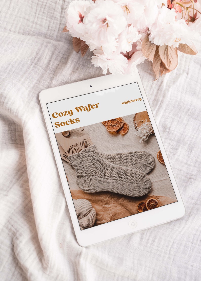 May include: A digital tablet displaying a knitting pattern for Cozy Wafer Socks by whileberry. The pattern features a close-up of the knitted socks on a brown background with dried orange slices.