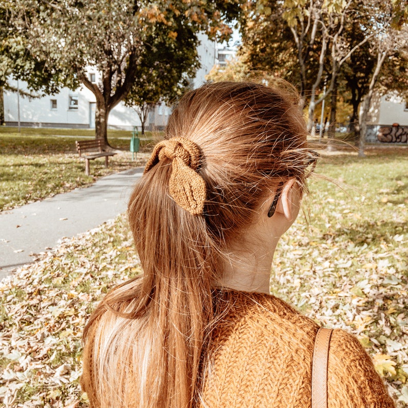 May include: A woman with long, brown hair wearing a brown sweater and a brown hair tie in the shape of a leaf. She is standing in a park with fallen leaves on the ground.