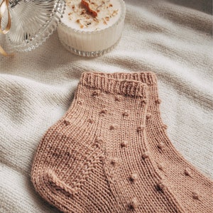 May include: A pair of knitted socks in a light brown color with a textured pattern of small dots. The socks are folded and displayed on a white fabric background.