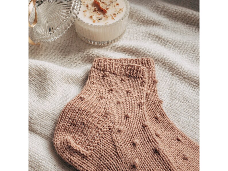 May include: A pair of knitted socks in a light brown color with a textured pattern of small dots. The socks are folded and displayed on a white fabric background.
