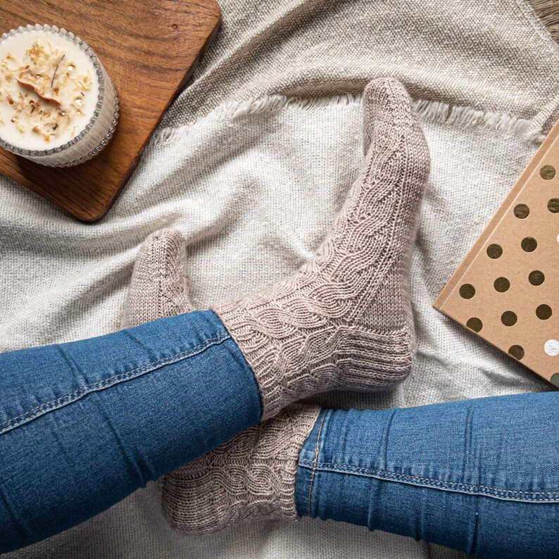 May include: A pair of beige cable-knit socks are worn with blue jeans. The socks have a textured pattern and are resting on a soft, light-colored blanket. A candle and a book are also visible.