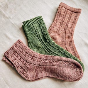 Peut inclure: Trois paires de chaussettes tricotées dans des tons de vert et de rose. Les chaussettes présentent divers motifs texturés, notamment des torsades et des côtes. Les chaussettes sont disposées sur une surface blanche texturée.