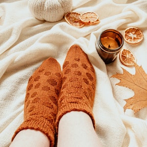 May include: A cozy autumn scene featuring a pair of orange patterned socks. A white knitted pumpkin, dried orange slices, a candle in a brown jar, and a fall leaf are arranged on a cream-colored blanket. The socks are the focal point.