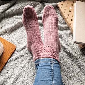 May include: A pair of knitted socks in a dusty rose color, featuring a ribbed design on the upper portion and a decorative lace pattern near the ankle. The socks are worn with blue jeans, resting on a gray, textured blanket.