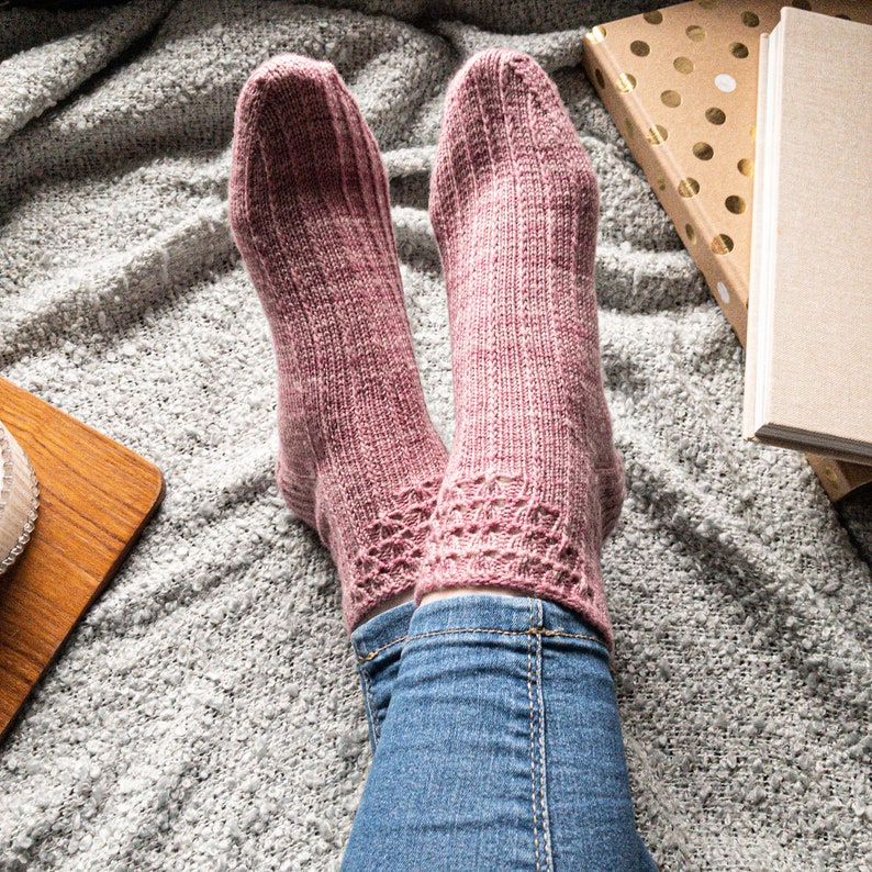 May include: A pair of knitted socks in a dusty rose color, featuring a ribbed design on the upper portion and a decorative lace pattern near the ankle. The socks are worn with blue jeans, resting on a gray, textured blanket.