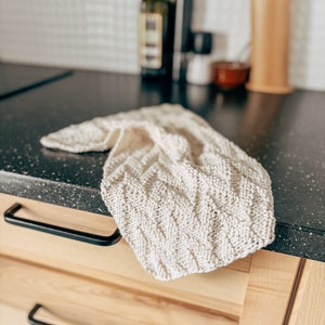 Puede incluir: Un paño de cocina de punto blanco con un patrón texturizado, doblado y colocado sobre una encimera de madera.