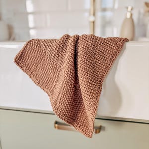 Op de afbeelding: Een vierkant, terracotta-kleurig washandje met een textuur van gebreide ribbelsteken. De stof is over de rand van een witte wastafel gevouwen en toont een consistent, dicht en uniform weefpatroon over het gehele oppervlak van het textiel.