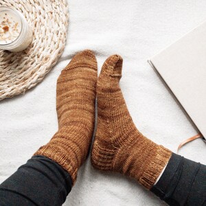 May include: A pair of brown knitted socks with a subtle marled pattern. The socks are worn on a person's feet and are resting on a white surface.