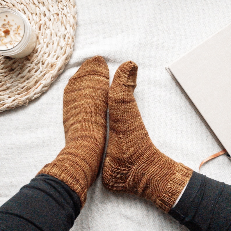 May include: A pair of brown knitted socks with a subtle marled pattern. The socks are worn on a person's feet and are resting on a white surface.