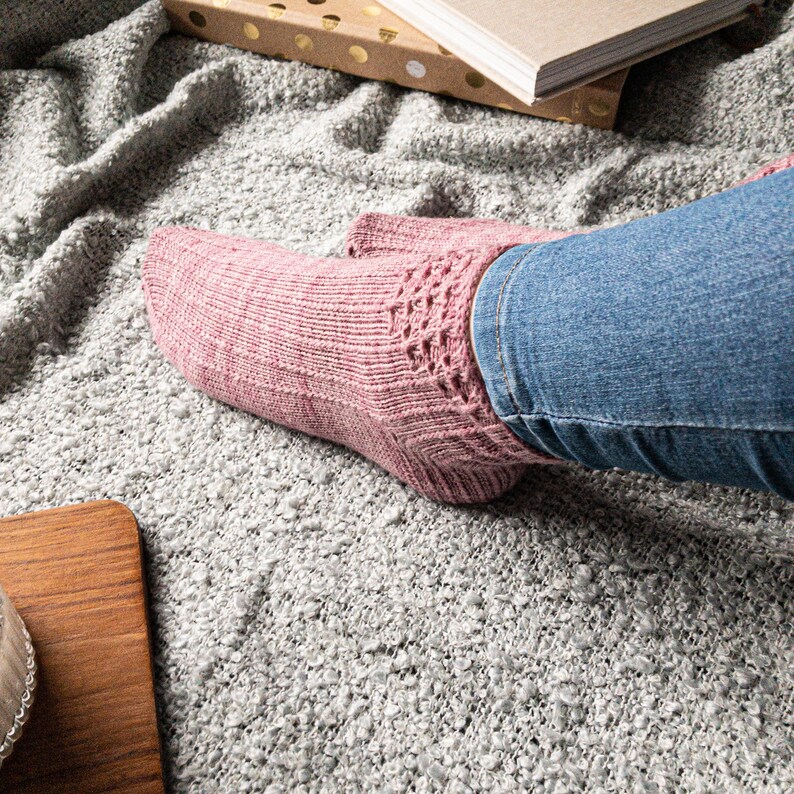 May include: A close-up of a pink knitted sock with a decorative openwork pattern on the ankle. The sock is worn with blue jeans. The background is a gray, textured blanket.